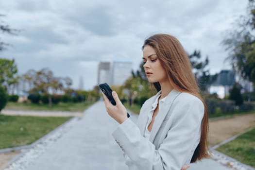 Thoughtful young woman in light blazer using smartphone outdoors in urban park during autumn, expressing focus and contemplation, business concept with neutral color palette.