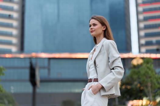 Confident business woman wearing light suit and white blouse stands outdoors near modern office buildings, expressing professionalism and determination in urban environment.
