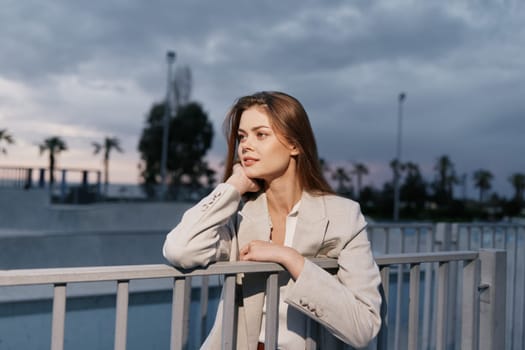 Thoughtful young woman wearing a light beige suit poses outdoors during sunset with a soft autumn urban background, showcasing casual fashion style and a confident woman mood.