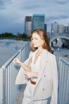 Confident young woman wearing beige suit walks on city bridge in cloudy weather, showcasing business fashion style with urban skyline. Woman expresses determination, professionalism and ambition.