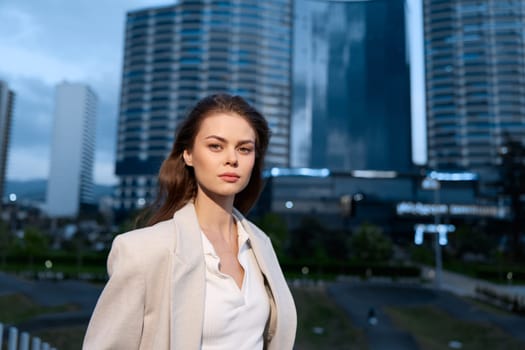 confident woman dressed in business casual attire stands outdoors near urban modern glass buildings at dusk, showing calm and professional expression in cool evening light