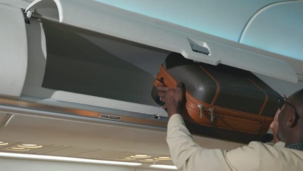 Passenger black man putting luggage on top shelf or cabin compartment on airplane, business man lift suitcases putting overhead locker on airplane, Travel concept