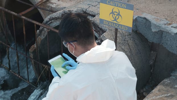 Ecologist sampling water of toxic chemicals from river touching screen on digital tablet, Biologist wear protective suit and mask collects sample of waste water from industrial, problem environment