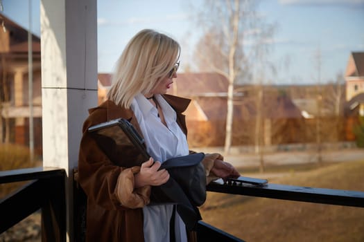 A woman with blonde hair stands on a balcony, dressed in a brown coat and sunglasses, holding a tablet in a tranquil neighborhood setting.