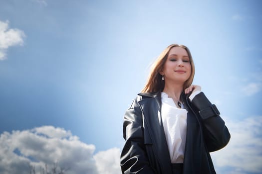 A young woman wearing a black coat stands with confidence under a clear sky, showcasing her style and poise.