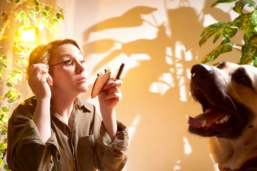A woman focuses on her makeup application as her dog sits close by, enjoying the warm sunlight in a plant-filled indoor space.