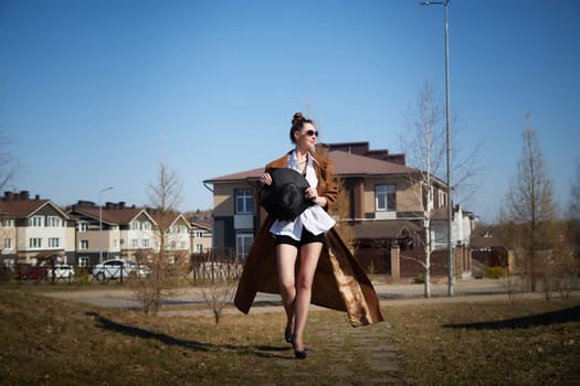 Model showcases a trendy ensemble featuring a flowing coat and a wide-brimmed hat while strolling through a neighborhood under blue skies.