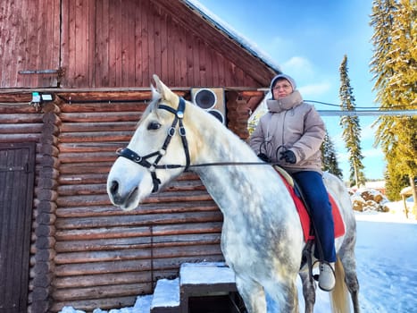 A person sits atop a white horse in a snowy environment beside a log cabin. The rider wears warm clothing, and the trees stand tall in the background under a clear sky.