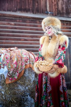 A girl in a vibrant national dress holds a basket of treats while posing with a horse during the Maslenitsa holiday.