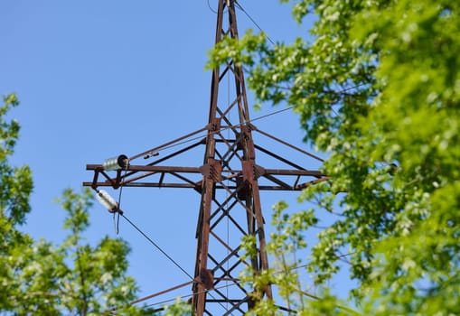 Upper part of an old, rusty power line tower against a blue sky and green tree leaves. Seen from a bottom-up perspective.