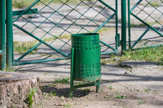 A green perforated metal trash can stands in a park or public area. Outdoor waste disposal bin