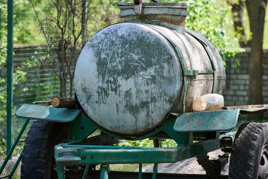 An old metal tank on a trailer, covered with traces of rust and time, stands against the background of green trees and a chain-link fence. The trailer is painted a worn green color