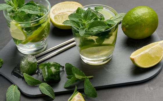 Glass of mojito cocktail with fresh mint leaves, lime slices, and ice cubes on a dark stone background. Refreshing summer drink with stainless steel straws