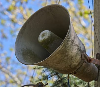 An old, dirty, grey loudspeaker is mounted on a pole. In the background, there is a blurred image of the sky and tree branches. View from below.