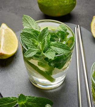 Glass of mojito cocktail with fresh mint leaves, lime slices, and ice cubes on a dark stone background. Refreshing summer drink with stainless steel straws