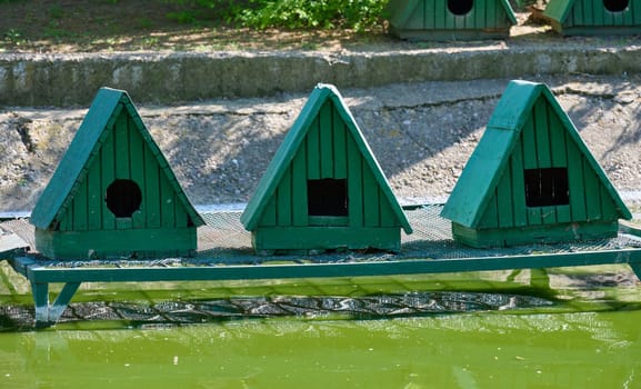 Small green wooden birdhouses with triangular roofs, set on a green metal platform over green water. Each birdhouse has an entrance hole