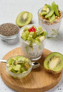 A glass of chia seed pudding layered with kiwi and granola, topped with kiwi slices, sits on a wooden board with ingredients around it