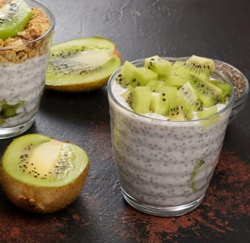 Two glasses of chia seed pudding, one topped with diced kiwi and the other with granola and banana chips, are on a dark surface with ingredients