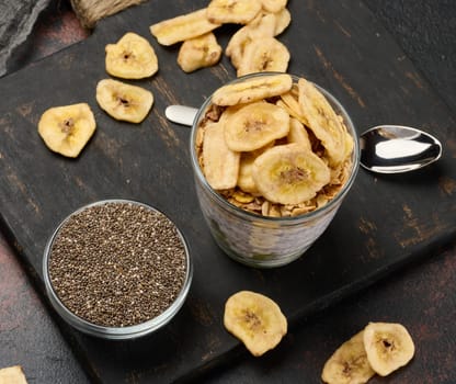 Glass of chia seed pudding layered with kiwi, topped with granola and banana chips, sits on a dark board with chia seeds and banana chips.
