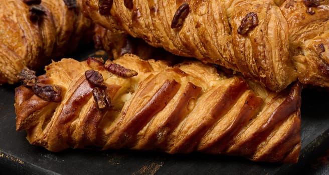 Golden brown maple pecan braid pastries are arranged on a dark, textured surface. Braided dough is flaky and topped with pecans