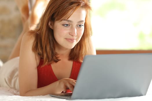 Satisfied woman using laptop lying on a bed in a house interior