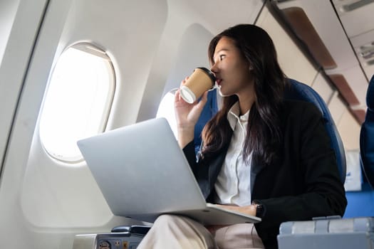 A businesswoman enjoying a coffee while using her laptop on an airplane, illustrating a modern approach to work during travel.