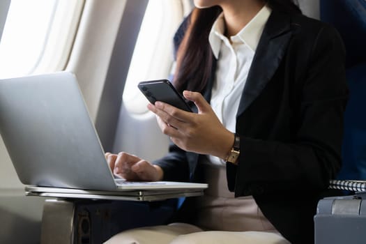 A young woman utilizes her smartphone and laptop simultaneously onboard a flight, emphasizing multitasking in business travel.