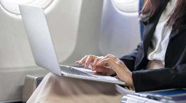 A close-up view of a young businesswoman's hands typing on a laptop while seated on an airplane, emphasizing productivity.