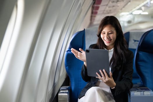 A young businesswoman joyfully waves at her tablet camera while seated on an airplane, connecting during her trip.