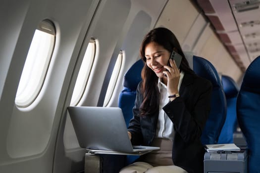 A young woman engaged in a phone call while using a laptop during her flight, highlighting productivity in business travel.