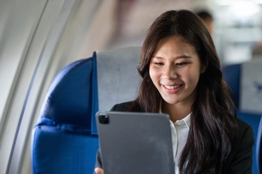 A young businesswoman smiles as she uses her tablet on an airplane, embracing productivity during her flight.