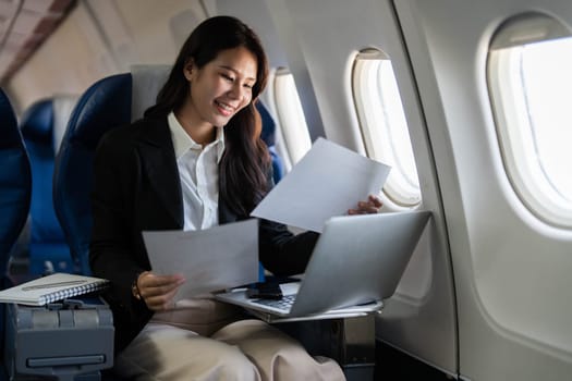 A happy professional woman smiling while reviewing documents and working on her laptop during a flight, representing positivity in business travel.