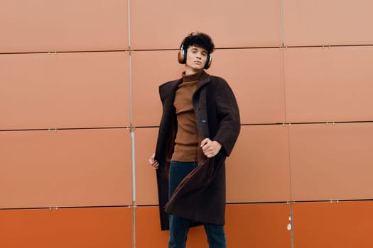 Fashionable young man in a coat and headphones standing against an orange wall with a smile on his face and hands in his pockets.