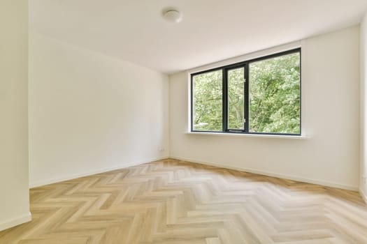 A bright and airy empty room featuring light wood flooring and a large window providing a view of greenery outside.