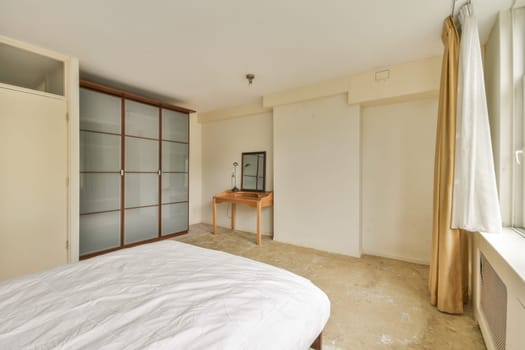 A spacious minimalist bedroom featuring a bed, a wooden table, a mirror, and large windows allowing natural light to flood the room. Neutral tones enhance serenity.