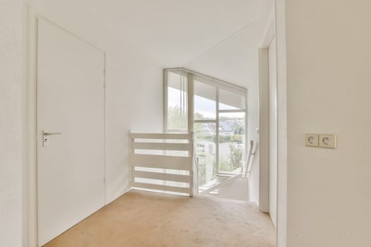 A spacious and well-lit interior hallway featuring a staircase with a modern design and large windows offering a view of the outdoors.