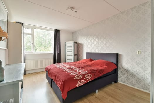 A stylish modern bedroom featuring a cozy red bedspread, wooden flooring, and large windows that bring in natural light.