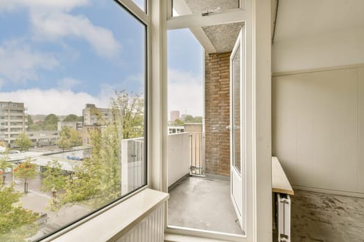 A modern interior view showcasing large windows, allowing natural light to flood a simple balcony space with urban greenery in sight.