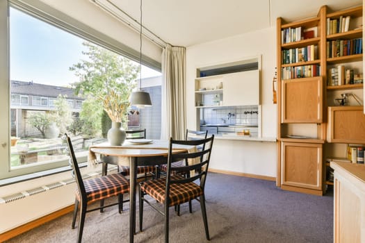 A well-lit dining space featuring a round table with checkered upholstery, surrounded by bookshelves and a view of greenery outside.