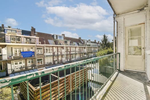 A scenic view showcasing a balcony with railings that overlooks a row of urban buildings under a blue sky with clouds. The greenery adds a touch of nature.