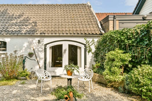 A cozy outdoor patio featuring white chairs and a small table surrounded by lush green plants. The inviting space is ideal for relaxation and leisure moments.