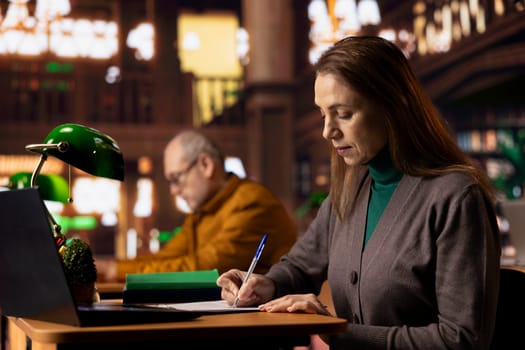 Mature female pupil writing class notes on clipboard files at the library, studying and reading at a college campus. Woman symbolizing preparation for her diploma thesis, intellect.