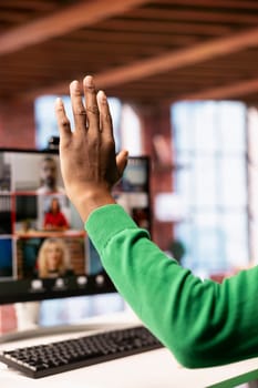 Teleworker at home office desk raising hand to request turn to speak during online videocall on PC. Close up of remote employee in apartment greeting team members during videoconference meeting