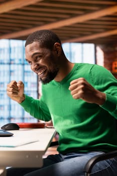 Overjoyed man thrilled by achievement, cheering and gesticulating at home. Happy african american person smiling, celebrating success, showing positive emotions, sitting at apartment desk