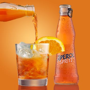Bottle and glass of aperol spritz cocktail with orange slice. The bartender pours aperol spritz into glass filled with ice cubes. Liquor on orange background.