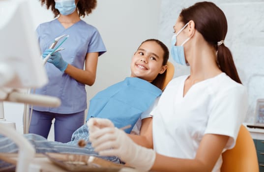 Work of medical dental team with small child. Millennial multiracial women in uniforms, protective masks and rubber gloves talk with smiling girl patient in medical chair in modern clinic, cropped
