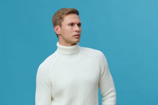 Male model in a white turtleneck sweater poses thoughtfully against a solid blue backdrop. The minimalistic studio setting highlights casual winter clothing, with a neutral expression and natural lighting conveying a calm, introspective mood. Concept: winter fashion