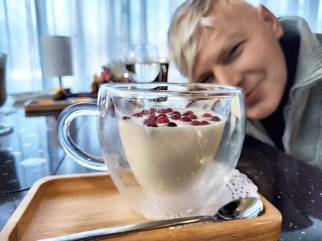 A man smiles in the background while a beautiful dessert rests on a wooden tray in the foreground. Colorful berries adorn the creamy treat, highlighting its freshness.