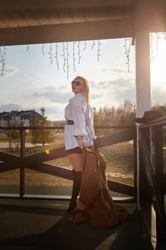 Beautiful girl with red hair in white shirt in open wooden pavillion in village or small town. Young slender woman and sky with clouds on background on autumn, spring or summer day
