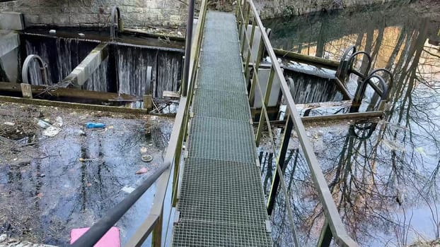A metal bridge spans calm water, surrounded by debris, showcasing industrial decay and environmental neglect